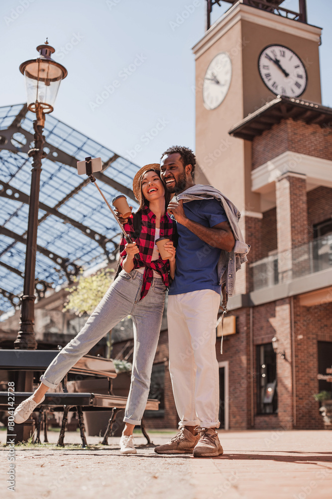 full length shot of happy asian girlfriend and african american boyfriend taking selfie on smartphone outdoors