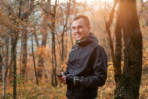 Concept of sport and active lifestyle. A young man in a black tracksuit, holding a mobile phone and smiling. In the background of autumn Park. Sunlight
