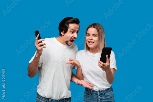Brunet man in white tees and blue jeans is shocked looking at the smartphone screen of a blond woman