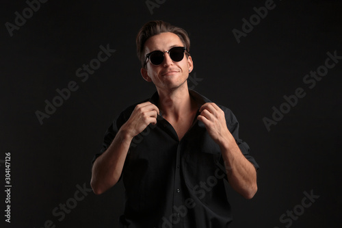 Stylish bussinesman in black eyeglasses buttoning up balck shirt isolated over black background