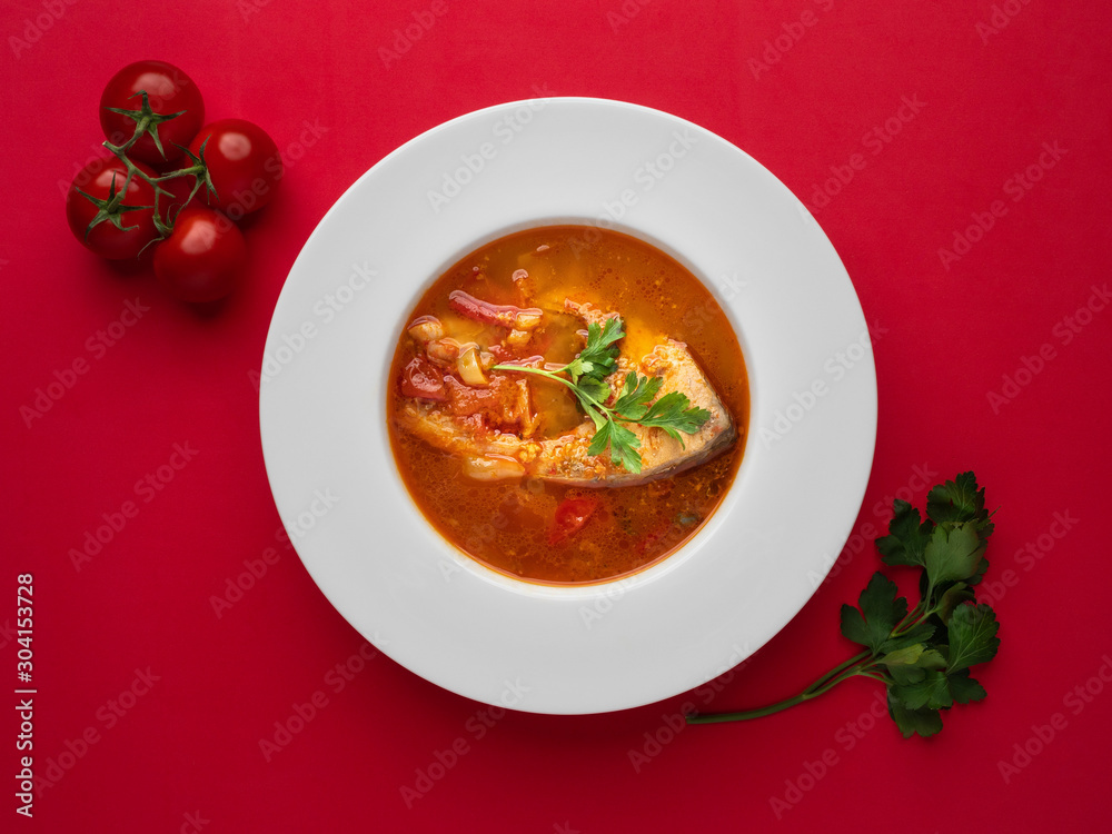 Fish soup in white plate with fresh tomatoes and parsley on red background. Overhead view. Fish soup in white plate with fresh tomatoes and parsley on red background. Overhead view.