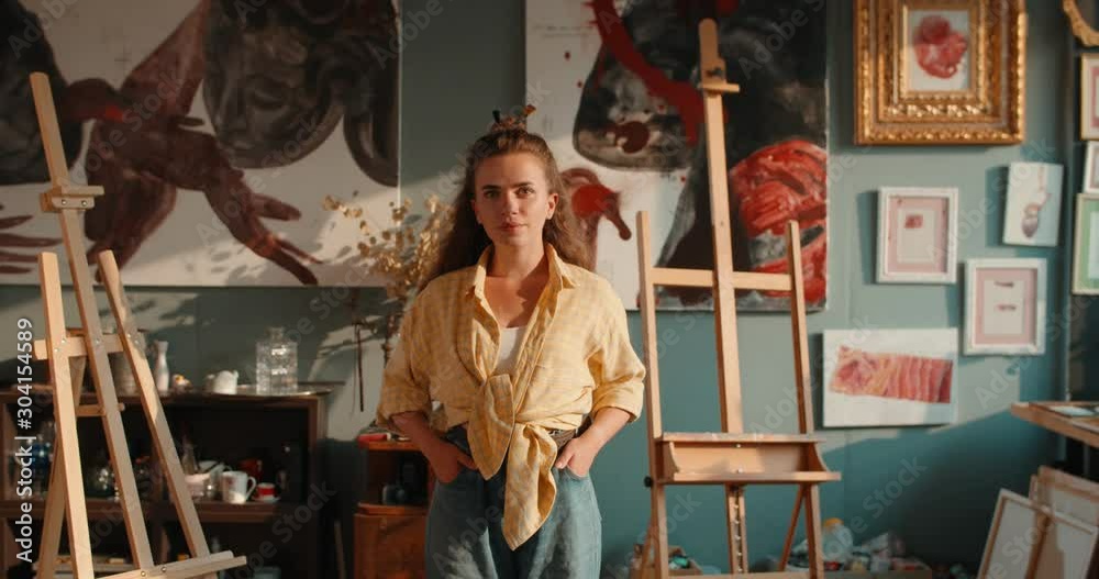 Portrait shot of the beautiful and cheerful young Caucasian woman artist standing in sunlight of her cozy art studio and smiling to the camera.