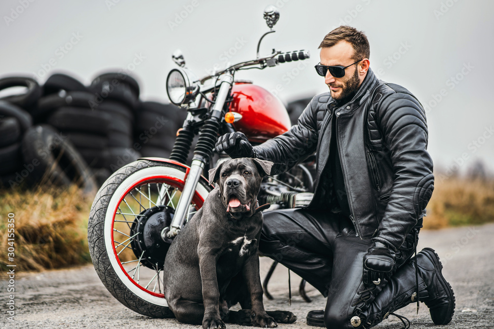 Biker in a leather suit crouched near his dog and red motorcycle on the road. Many tires on the background