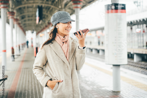 Young woman in winter coat and hats with phone in hand waiting on railway station for train. Happy young smiling girl standing in the ststion while making voice messages. photo