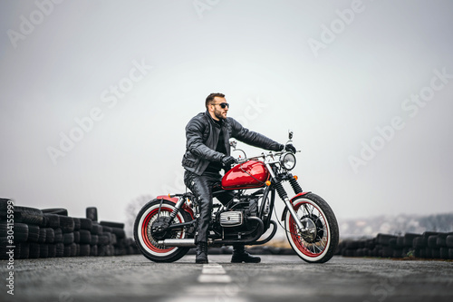 Red motorbike with rider. A man in a black leather jacket and pants stands sideways in the middle of the road. Tires are laid on the background