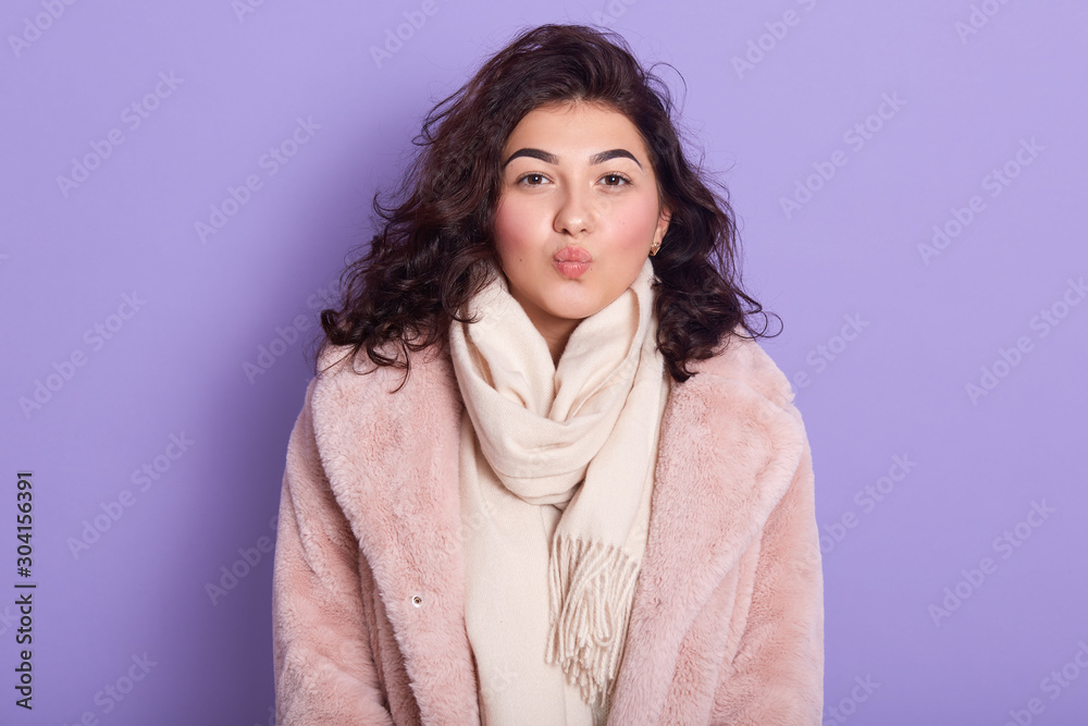 Portrait of beautiful girl dresses winter faux fur coat looking at camera and making kiss gesture isolated over lilac studio background, female wears only eco furs, lady with dark wavy hair. Winter. Portrait of beautiful girl dresses winter faux fur coat looking at camera and making kiss gesture isolated over lilac studio background, female wears only eco furs, lady with dark wavy hair. Winter.
