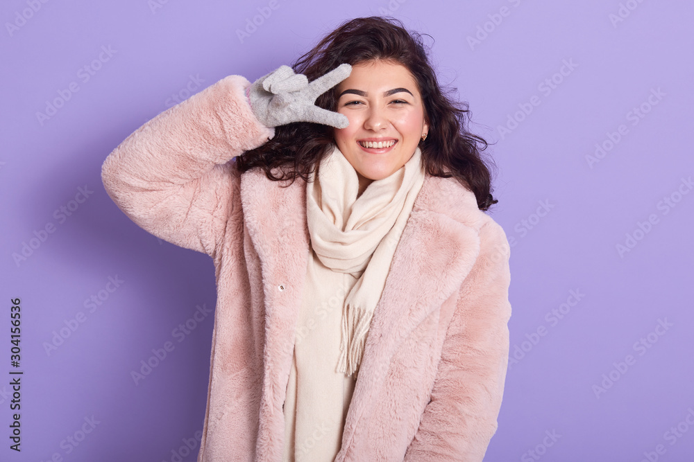 Cheerful girl with dark wavy hair posing with peace sign, adorable female wearing pale pink faux fur coat, scarf and gloves, caucasian woman having fun in winter, covering her eye with v sign. Cheerful girl with dark wavy hair posing with peace sign, adorable female wearing pale pink faux fur coat, scarf and gloves, caucasian woman having fun in winter, covering her eye with v sign.