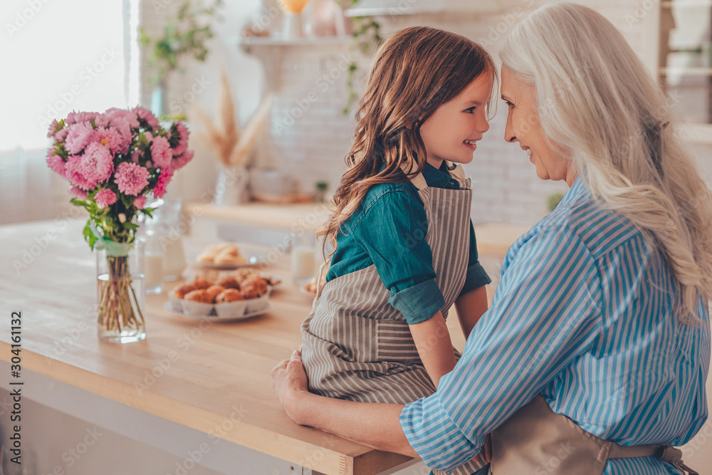 little girl sitting on the table and grandmother standing on the kitchen and looking at each other, side view little girl sitting on the table and grandmother standing on the kitchen and looking at each other, side view