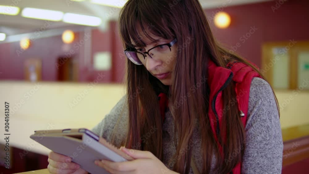 Portrait of young woman with glasses uses tablet computer in mall. Brunette girl is surfing and browsing online, mobile office for work or training. Portrait of young woman with glasses uses tablet computer in mall. Brunette girl is surfing and browsing online, mobile office for work or training.