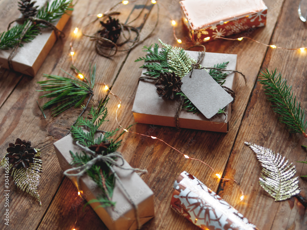 Christmas presents wrapped in craft paper with natural fir tree and thuja branches as decoration. Wooden table with light bulbs and hand made New Year gifts. Christmas presents wrapped in craft paper with natural fir tree and thuja branches as decoration. Wooden table with light bulbs and hand made New Year gifts.