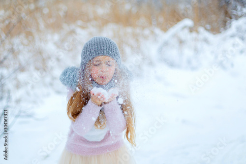 Pretty little girl wearing grey knitted hat and pink sweater walking in snowy park.  Cute child blowing snowflakes in winter forest. Family vacation with kid on christmas holidays. Blurred background