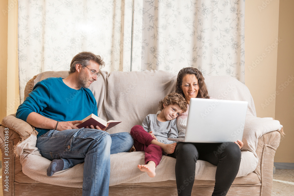 Child, a woman and a man sitting on a sofa watching a laptop and reading a book. Child, a woman and a man sitting on a sofa watching a laptop and reading a book.