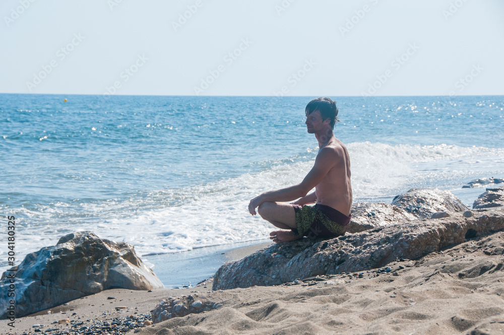 Young man doing yoga on a beach. Man sitting on a rock by the sea. Summer vacation. Mediterranean Sea.