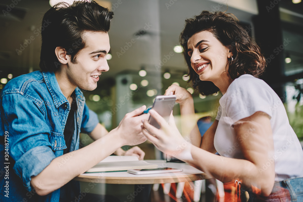 Happy smiling man and woman watching funny video online on content website via cellular phone, positive caucasian friends enjoying communication via smartphone gadget during free time in cafeteria