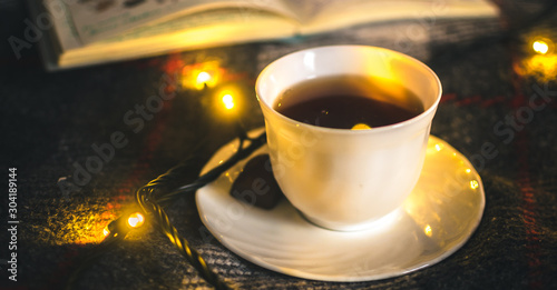 A cup of hot black tea with chocolate on a saucer on a warm and cozy plaid with Christmas lights
