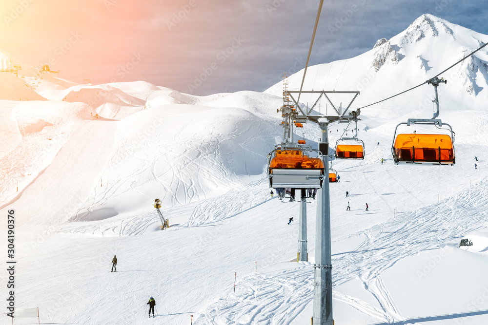 Ski lift ropeway on hilghland alpine mountain winter resort on bright sunny day. Ski chairlift cable way with people enjoy skiing and snowboarding.Banner panoramic wide view of downhill slopes Ski lift ropeway on hilghland alpine mountain winter resort on bright sunny day. Ski chairlift cable way with people enjoy skiing and snowboarding.Banner panoramic wide view of downhill slopes