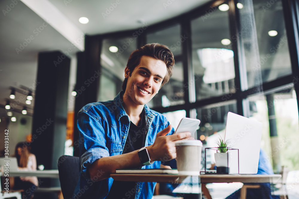 Portrait of cheerful caucasian young man smiling at camera while holding smartphone in hands and updating profile of blog sitting at laptop, generation of millennial guy using modern devices Portrait of cheerful caucasian young man smiling at camera while holding smartphone in hands and updating profile of blog sitting at laptop, generation of millennial guy using modern devices