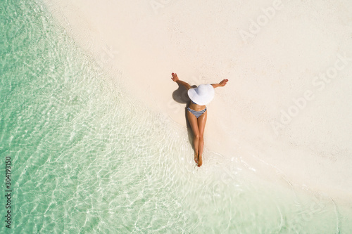 Summer holiday fashion concept - tanning girl wearing sun hat at the beach on a white sand shot from above.Top view from drone. Aerial view of slim woman sunbathing lying on a beach in Maldives. photo