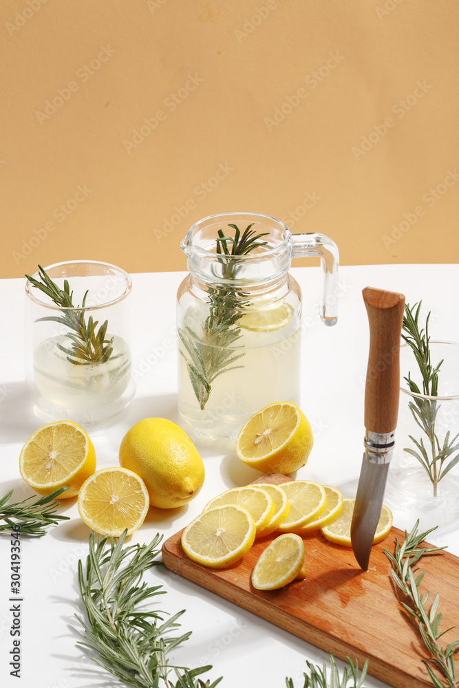 Jug with homemade lemonade, glasses with ice, lemons and rosemary on a white table.