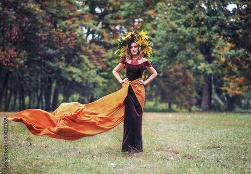 Portrait of beautiful girl with curly balayage hair and autumn wreath on the head wearing velvet burgundy dress and orange fabric posing in the wood