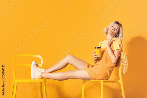 Fashionable young woman with coffee sitting on chair near color wall
