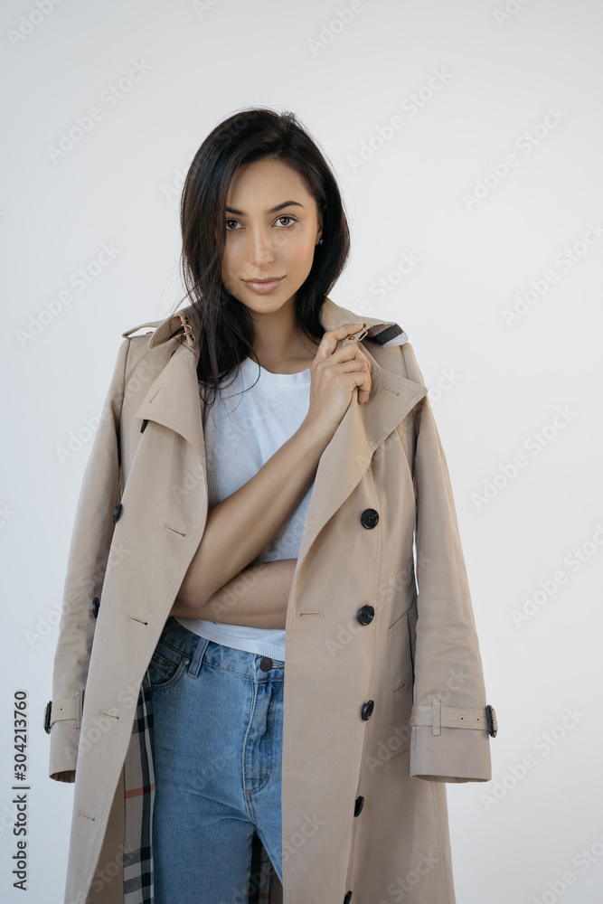 Attractive asian woman wearing stylish trench coat isolated on white background. Successful Indian fashion model with beautiful face and body posing for pictures in studio, looking at camera Attractive asian woman wearing stylish trench coat isolated on white background. Successful Indian fashion model with beautiful face and body posing for pictures in studio, looking at camera