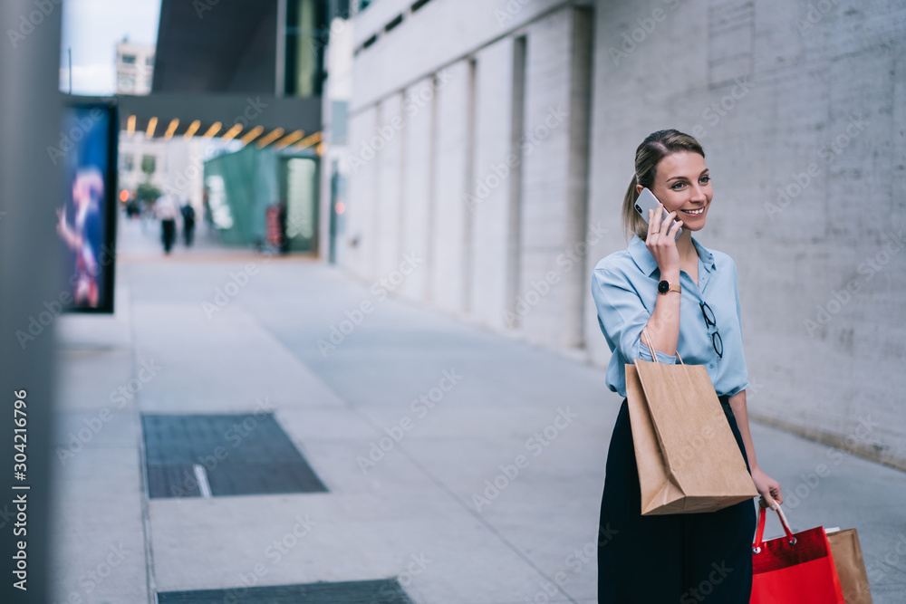 Fototapeta premium Cool cheerful hipster girl in stylish wear talking on smartphone in roaming, happy young woman calling on mobile phone device standing on street in modern city with paper bags during Black Friday