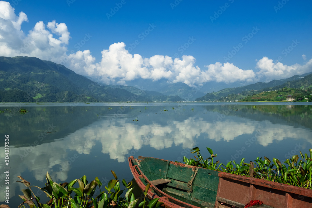 View of the lake with turquoise water and the opposite mountain shore. Reflection of clouds in calm water. Phewa Lake in Pokhara (Nepal) in the morning