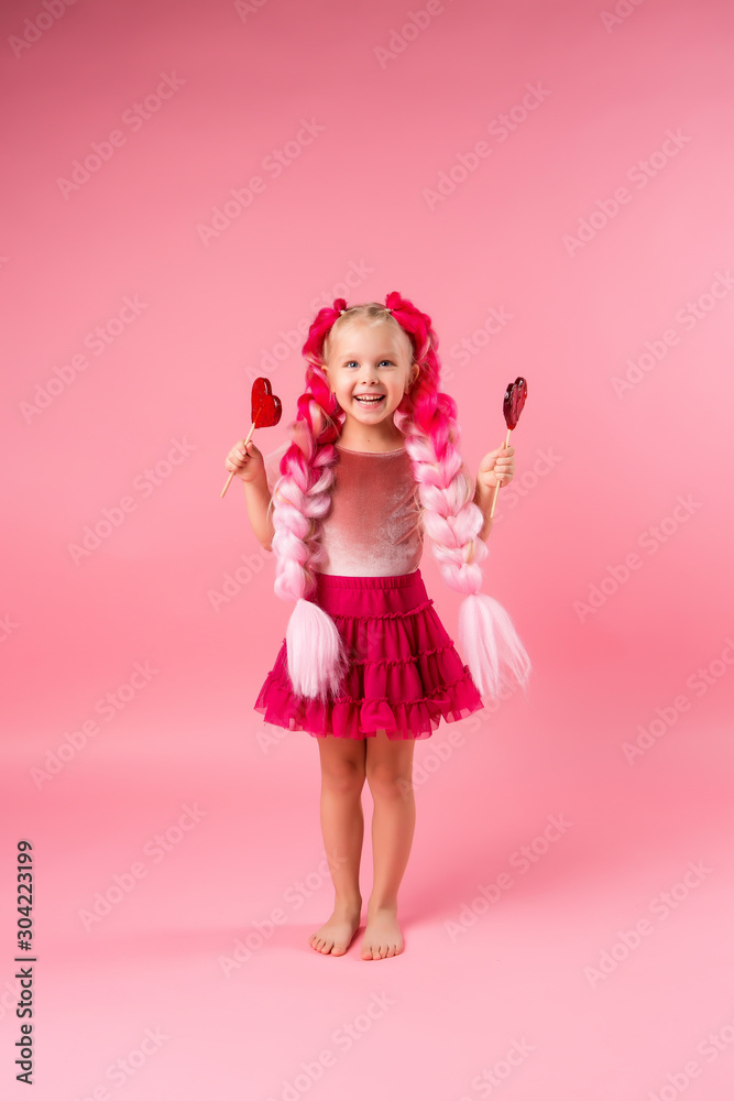 cute little girl in a pink dress with pink braids from kanekalon holds heart-shaped lollipops in her hands cute little girl in a pink dress with pink braids from kanekalon holds heart-shaped lollipops in her hands