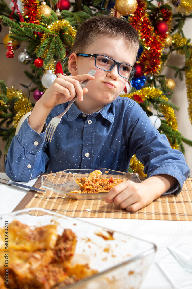 Chłopiec w okularach siedzi przy stole i ma buzię pełną jedzenia. Na talerzu leży lasagne a w tle pięknie ubrana choinka. Święta Bożego Narodzenia jedzenie. - obrazy, fototapety, plakaty 