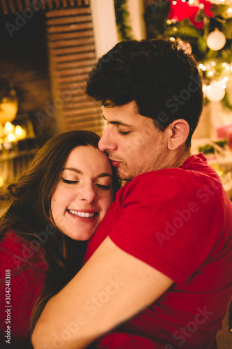 Couple celebrating Christmas together. Attractive woman and handsome man with close eyes hugging under the fireplace with candles, lights, presents and decorated tree. Happy New Year concept.