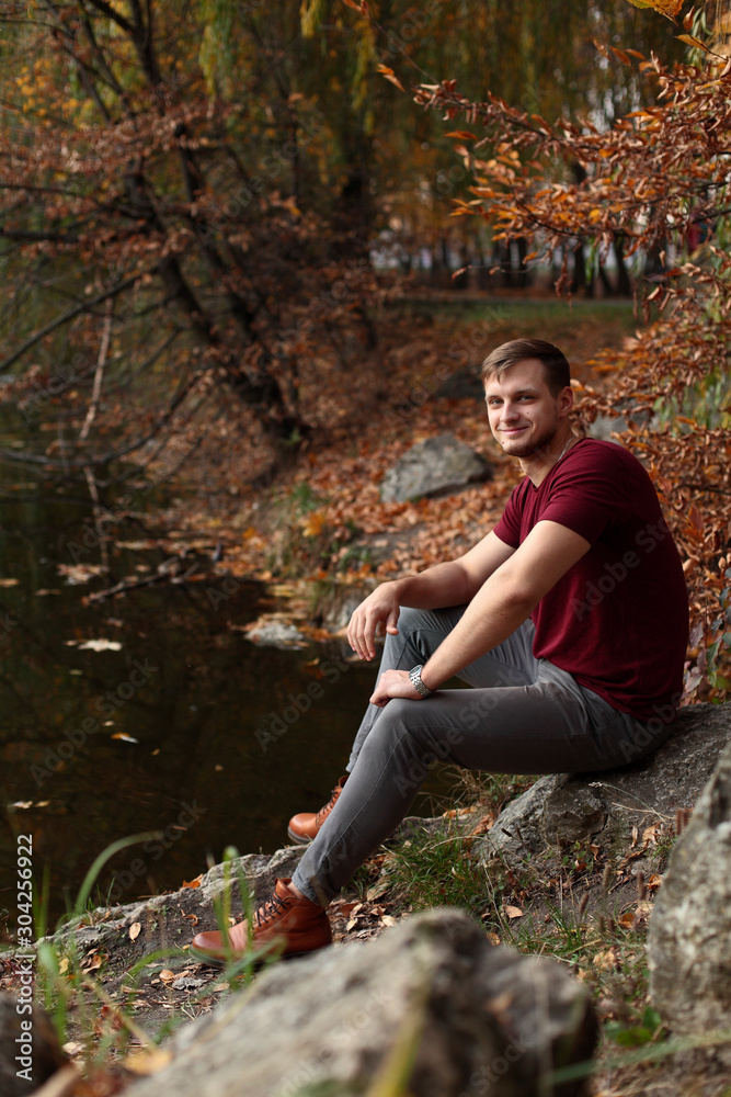A man sits on the bank of the river in the autumn and looks into the camera, a photo in warm colors