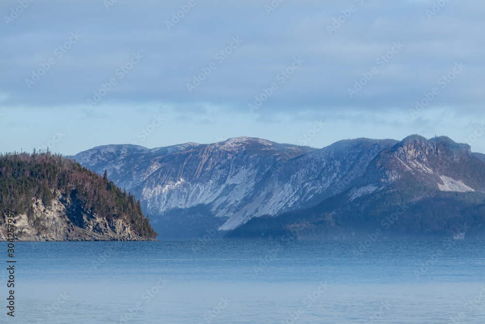 Snow and trees covered mountains in the background under thick clouds and blue sky. The ocean is in the foreground with soft reflections. There's texture throughout the photo.