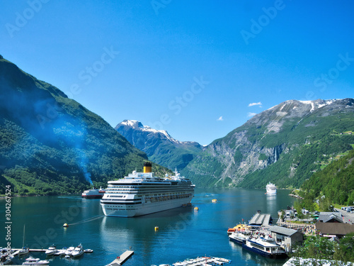 Spectacular view of Geiranger Fjord and mountains with snow, waterfalls, moored cruise ships and tender boats carrying tourists from ship to shore on a beautiful day with blue sky.