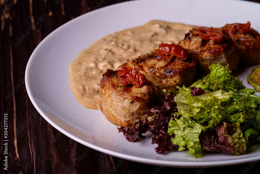Pork medallions with vegetables on white plate, wooden background, dark light, angle view