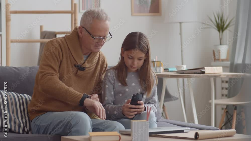 Medium shot of Caucasian pretty girl sitting on sofa together with her grandfather and helping him mastering mobile phone and smart watch on his hand Medium shot of Caucasian pretty girl sitting on sofa together with her grandfather and helping him mastering mobile phone and smart watch on his hand