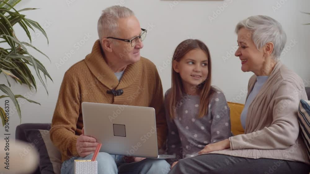 Medium shot of Caucasian grandparents and their cute granddaughter sitting on couch and using laptop together Medium shot of Caucasian grandparents and their cute granddaughter sitting on couch and using laptop together
