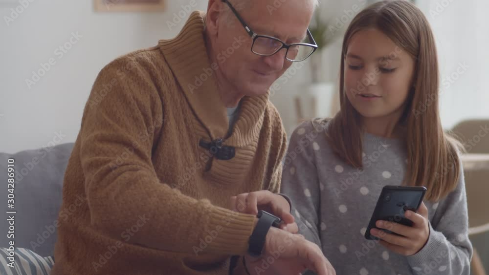 Medium shot of Caucasian beautiful girl sitting on couch in living room with her grandfather and helping him mastering cellphone and smart watch on his wrist