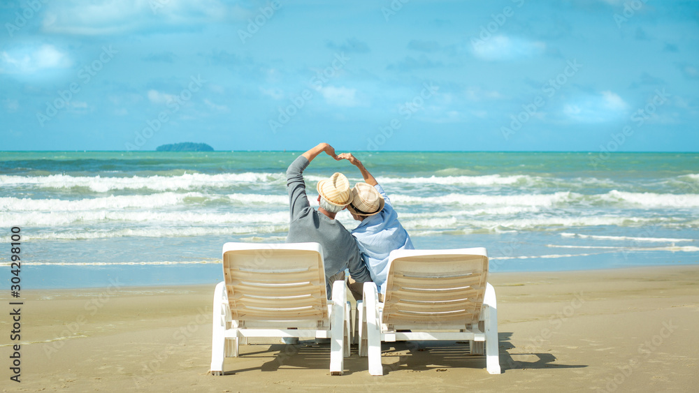 Rear view of senior couple sitting in deckchair on beach and Love making heart with hands. Love is everything, Retirement age concept and love