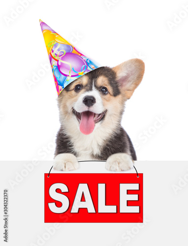 Corgi puppy in birthday hat holds sales symbol above white banner. isolated on white background