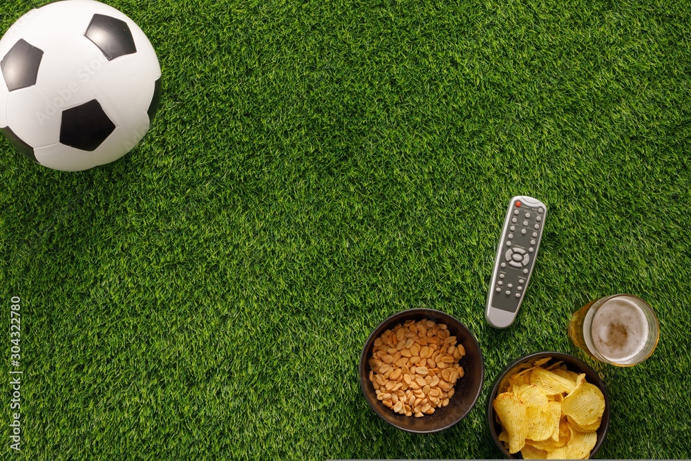 Naklejka premium Soccer ball on a green field and ottoman for a fan with snacks and a TV remote control. flat lay.Copy space.