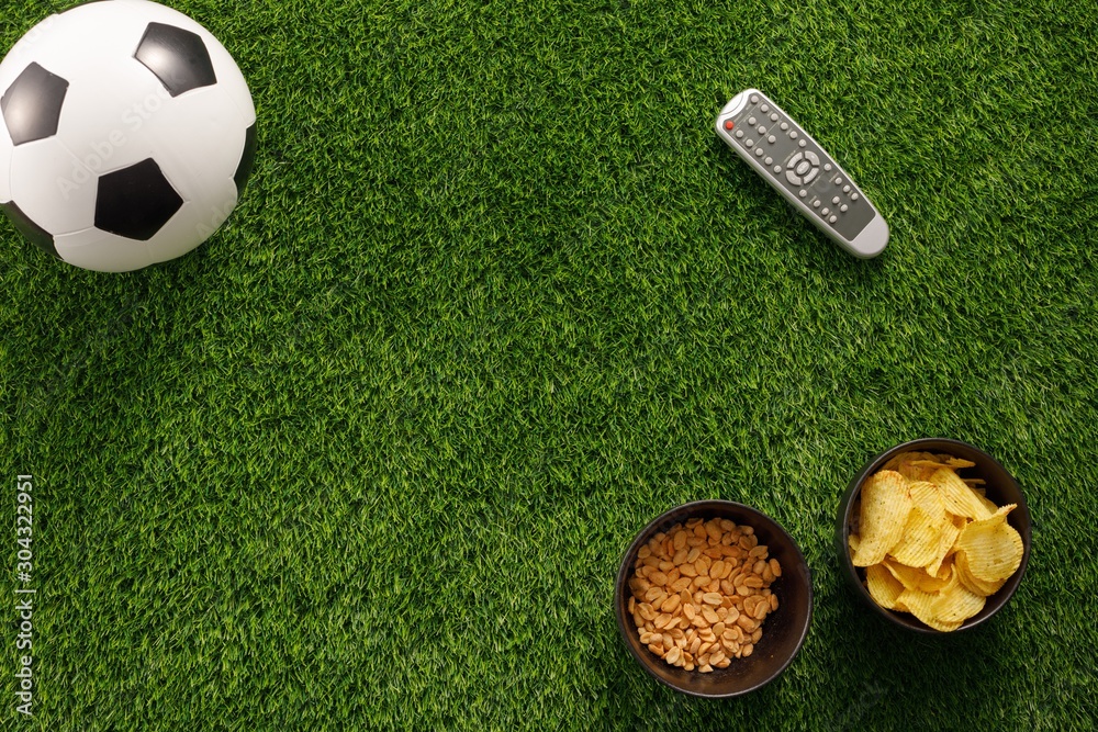 Naklejka premium Soccer ball on a green field and ottoman for a fan with snacks and a TV remote control. flat lay.Copy space.