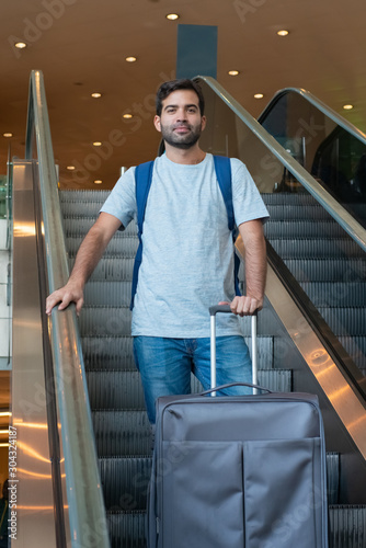 Cheerful traveler with suitcase on escalator. Cheerful traveler looking at camera. Travel concept