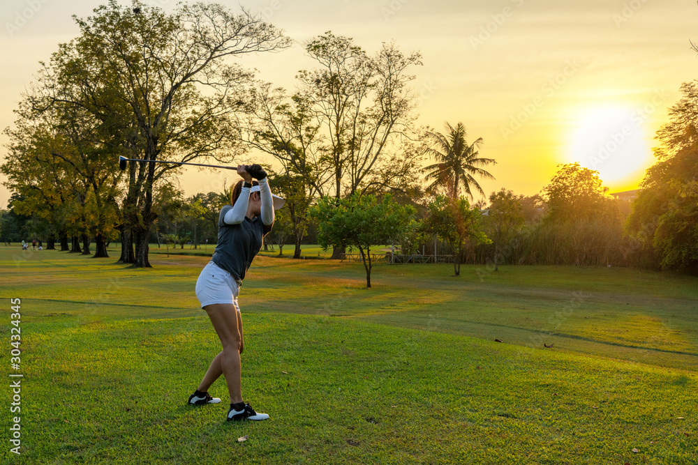 Healthy Sport. Asian Sporty woman golfer player doing golf swing tee off on the green course evening time, she presumably does exercise outdoor.  Healthy Lifestyle Concept..