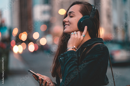 Young happy stylish trendy casual hipster woman teenager listening to music on a black wireless headphone while walking around the city. Music lover enjoying music photo