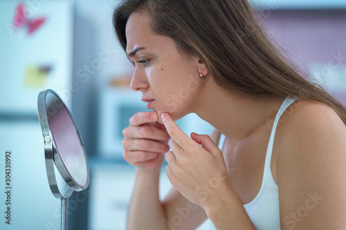 Upset stressed sad acne woman with problem skin squeezes pimple at home in front of a small round mirror