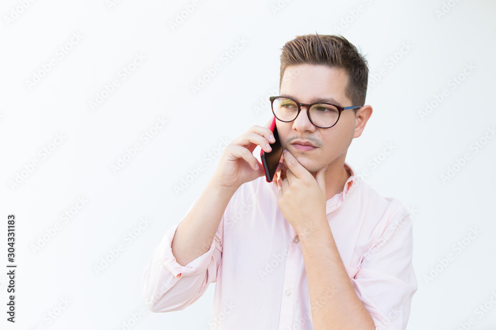 Serious pensive professional getting offer on cell, touching chin and thinking. Young man in glasses standing isolated over white background. Phone talk concept Serious pensive professional getting offer on cell, touching chin and thinking. Young man in glasses standing isolated over white background. Phone talk concept