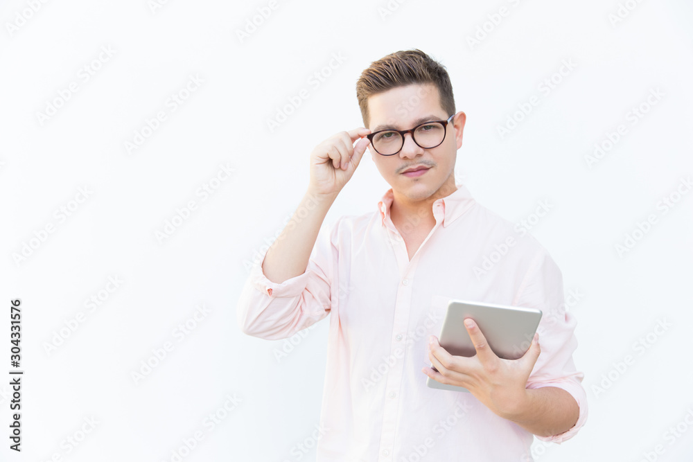 Serious confident app developer holding tablet, adjusting glasses, looking at camera. Young man in glasses standing isolated over white background. Software developer concept Serious confident app developer holding tablet, adjusting glasses, looking at camera. Young man in glasses standing isolated over white background. Software developer concept