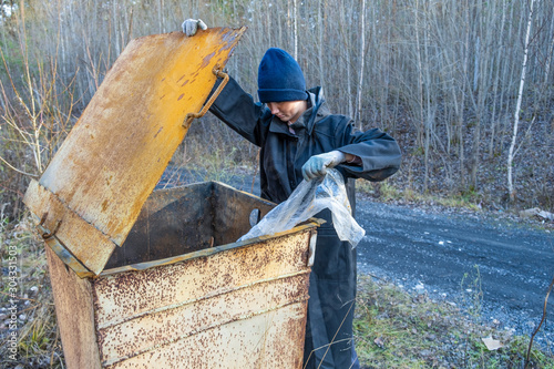 homeless woman in black coat and blue hat is open street garbage cans. woman is rummaging for some food in garbage. poverty.