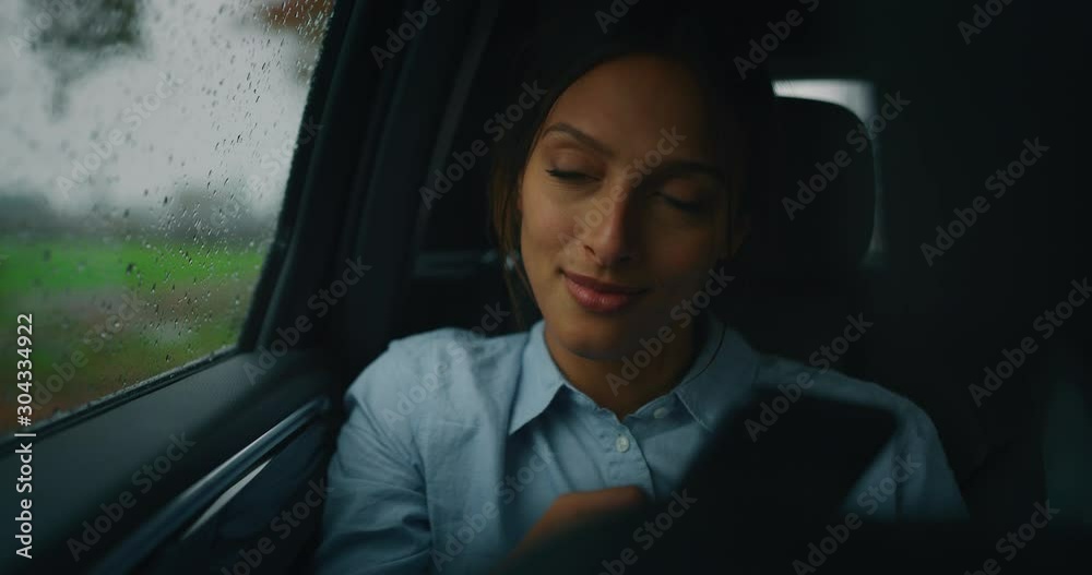 An young businesswoman is using a smartphone during traveling in a modern car with a driver while is raining outside. An young businesswoman is using a smartphone during traveling in a modern car with a driver while is raining outside.